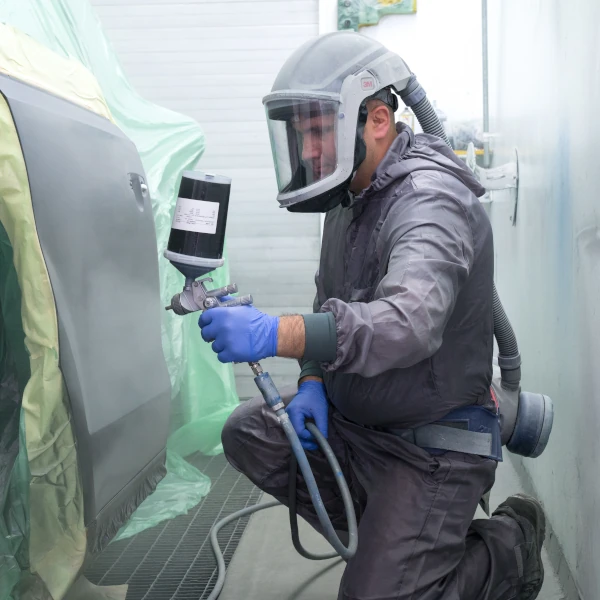 Kfz-Lackierer trägt Schutzanzug und lackiert Autotür in moderner Lackierkabine.