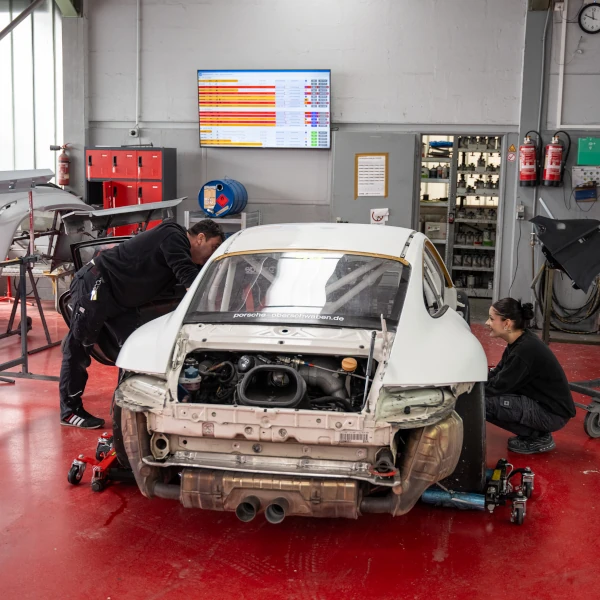 Zwei Mechaniker arbeiten in einer Autowerkstatt an einem weißen Sportwagen ohne Heckstoßstange.